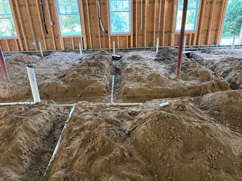 Open trenches cut across the dirt floor with white PVC drain lines laid in them and string lines stretched overhead.