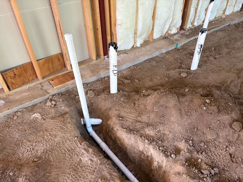 Close-up of capped PVC stub-ups hand-labelled in marker beside a trench with a drain line.
